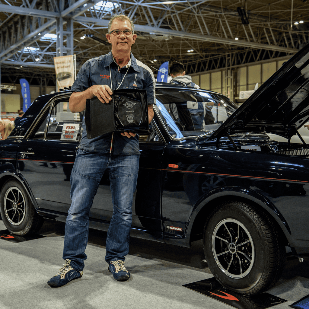 A proud man is standing next to a dark blue Ford Cortina Savage at an indoor motor show. He is holding a trophy and a plaque, with a smile on his face. The classic car's hood is open, revealing its engine. The vehicle is on a blue carpet with a large Ford logo. Other classic cars and attendees are visible in the background, creating a bustling exhibition environment.