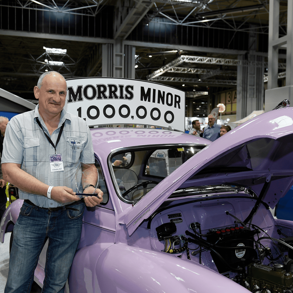 A man in a checkered shirt and jeans is standing next to a light purple Morris Minor with its hood open at a car show. He is holding a pair of glasses in his hands and is looking directly at the camera. A large sign in the background reads 