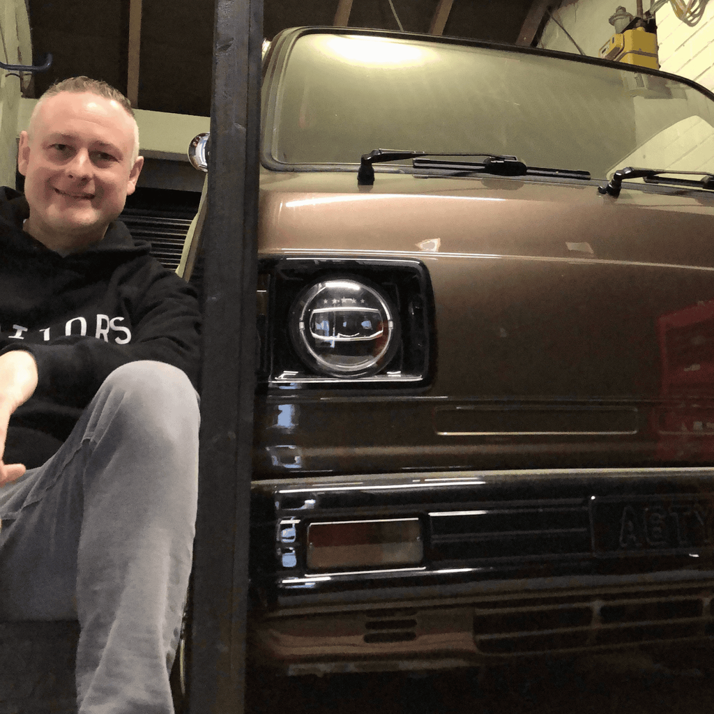 A man is sitting next to a custom brown Honda Acty TN550 Kei truck inside a garage or workshop. He is smiling at the camera and is wearing a black hoodie with a white logo. The front of the vintage Japanese truck, which is the main subject, is clearly visible, showing its lowered stance and modern, custom headlights. The license plate 