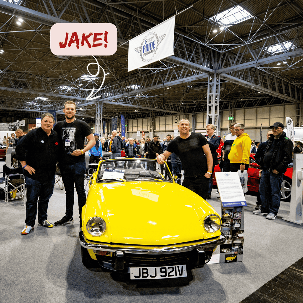 A group of people are posing with a bright yellow Triumph Spitfire convertible at an indoor car show. The car, which is the main subject, has its soft top down and a license plate that reads 