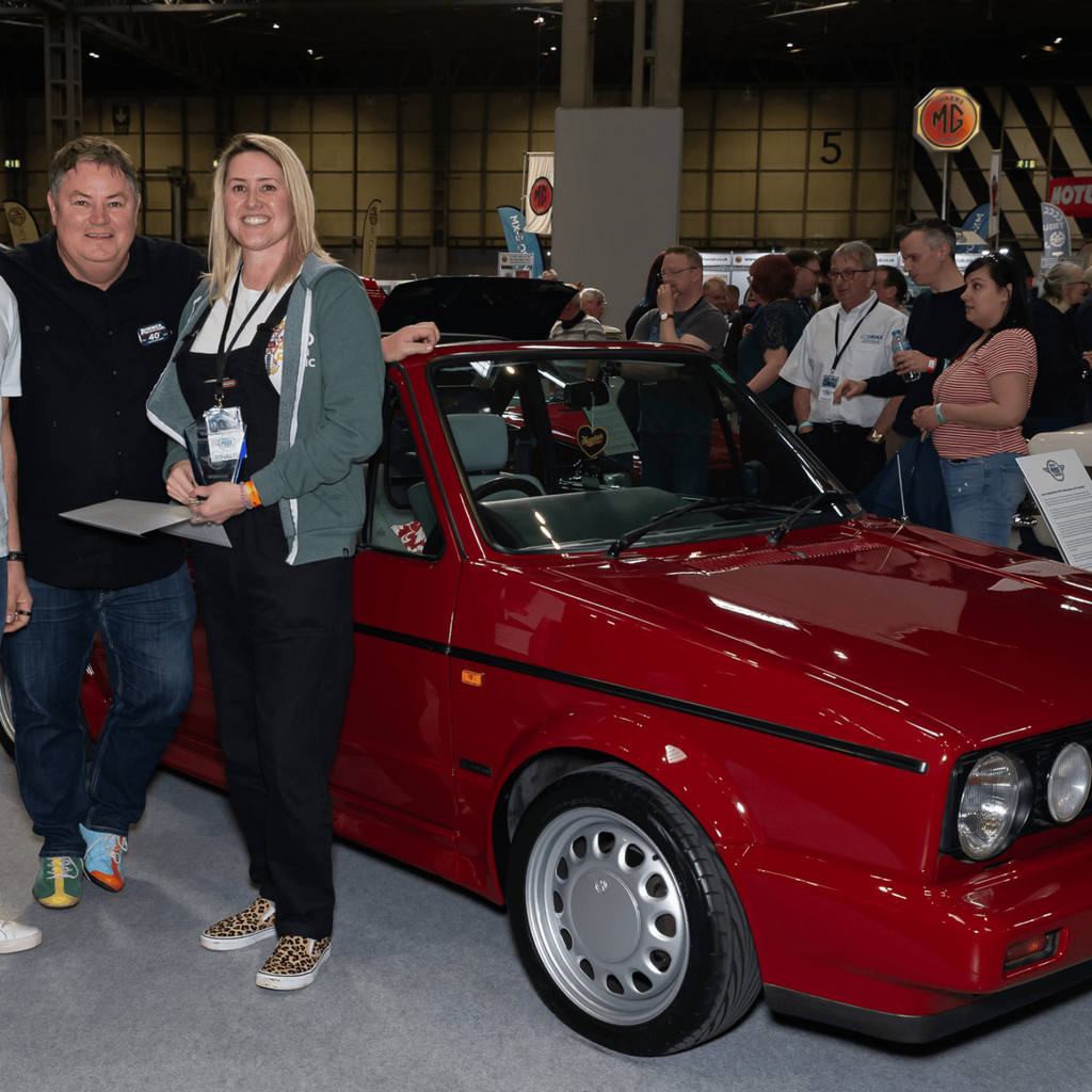 A woman is standing proudly next to a red Volkswagen Golf GTI Cabriolet convertible at an indoor car show, holding a framed certificate. Two men are standing with her, with the group posed next to the car. The car's license plate 