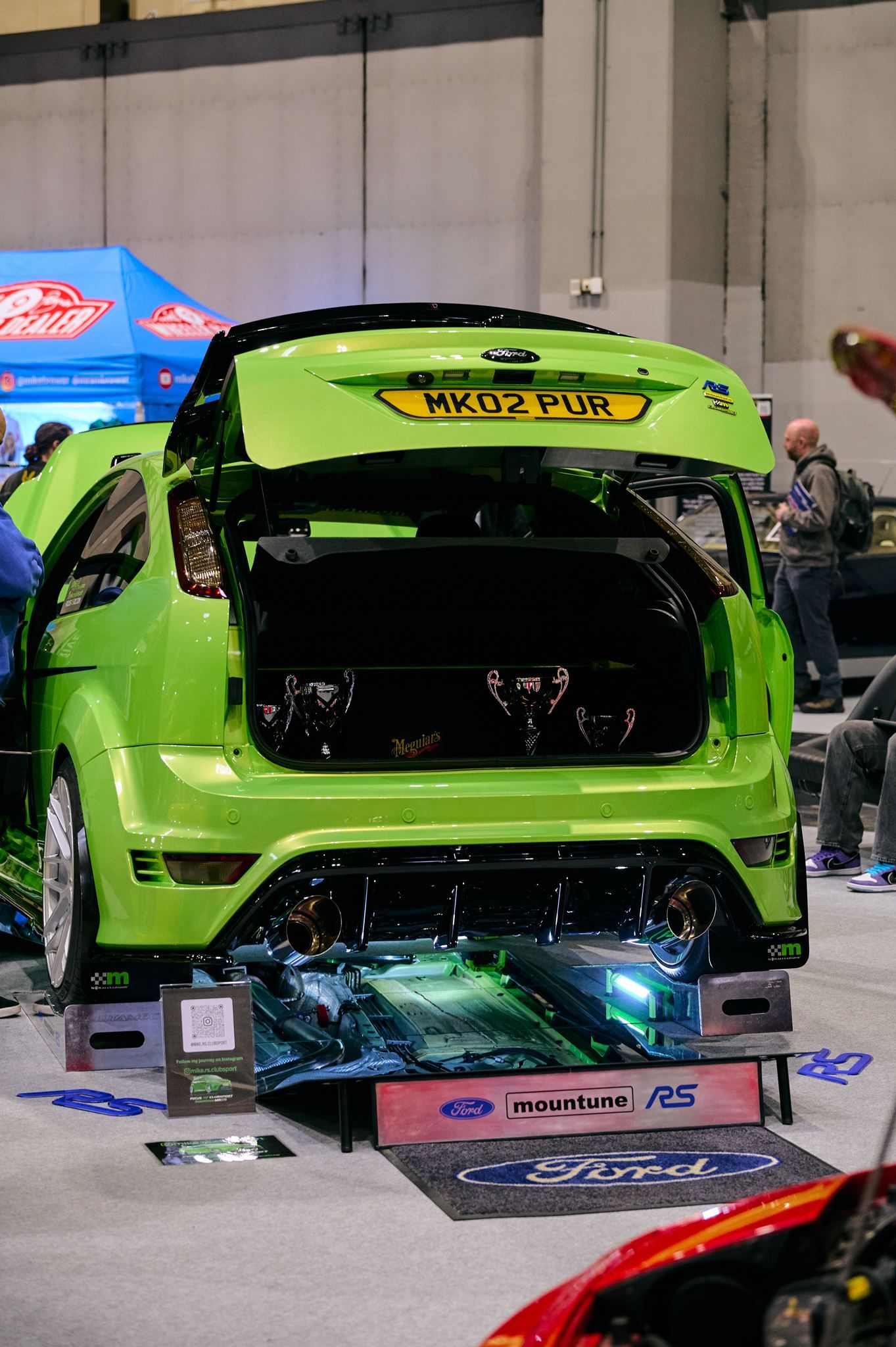 Rear view of a bright green Ford Focus RS on display with open boot, custom exhaust, underbody lighting, and trophies.