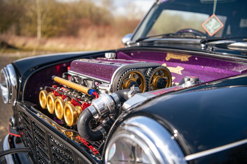 Close-up of a classic Mini engine bay featuring a purple metallic valve cover, gold velocity stacks, and chrome performance radiator details.