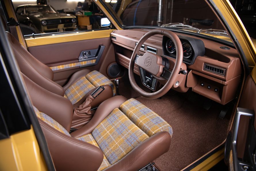 Custom interior of a restored yellow Austin Allegro featuring brown leather bucket seats with plaid fabric inserts and a leather-wrapped steering wheel.