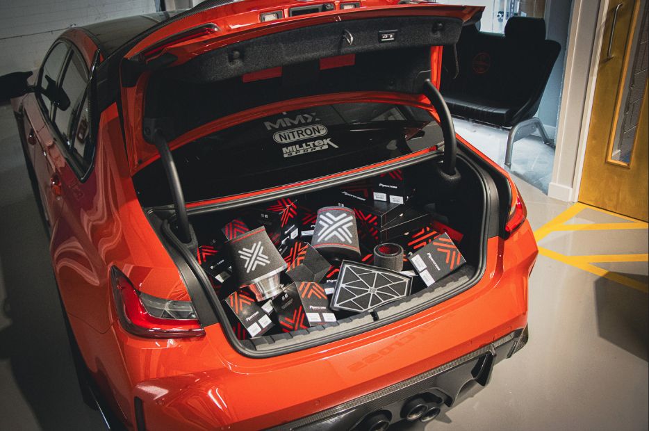 The open boot of a bright orange performance sedan, possibly a BMW M3, filled with a large collection of black and red aftermarket car products, boxes, and air filters.