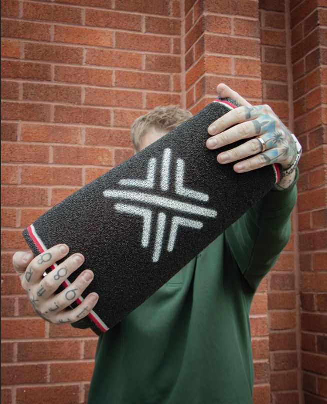 Man with tattooed hands and a green sweatshirt holding a rolled-up black aftermarket air filter or foam element with a white cross logo against a red brick background.