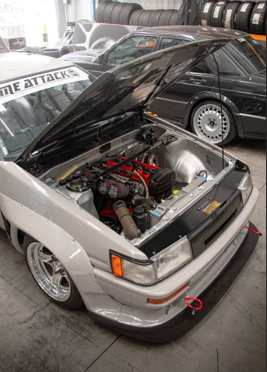 Highly modified grey classic JDM Toyota Corolla AE86 with a bright red 4AGE engine swap and wide body kit, bonnet open in a workshop next to a dark Mercedes-Benz.