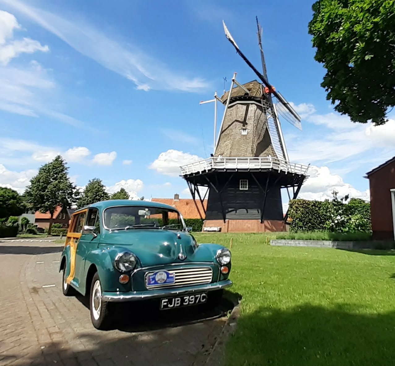A vintage green Morris Minor Traveller with timber frame trim and license plate FJB 397C parked on a paved lane in front of a tall traditional windmill under a blue sky.