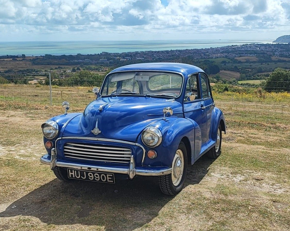 A classic bright blue Morris Minor saloon with license plate HUJ 990E parked on a grassy hilltop overlooking a scenic coastal bay.