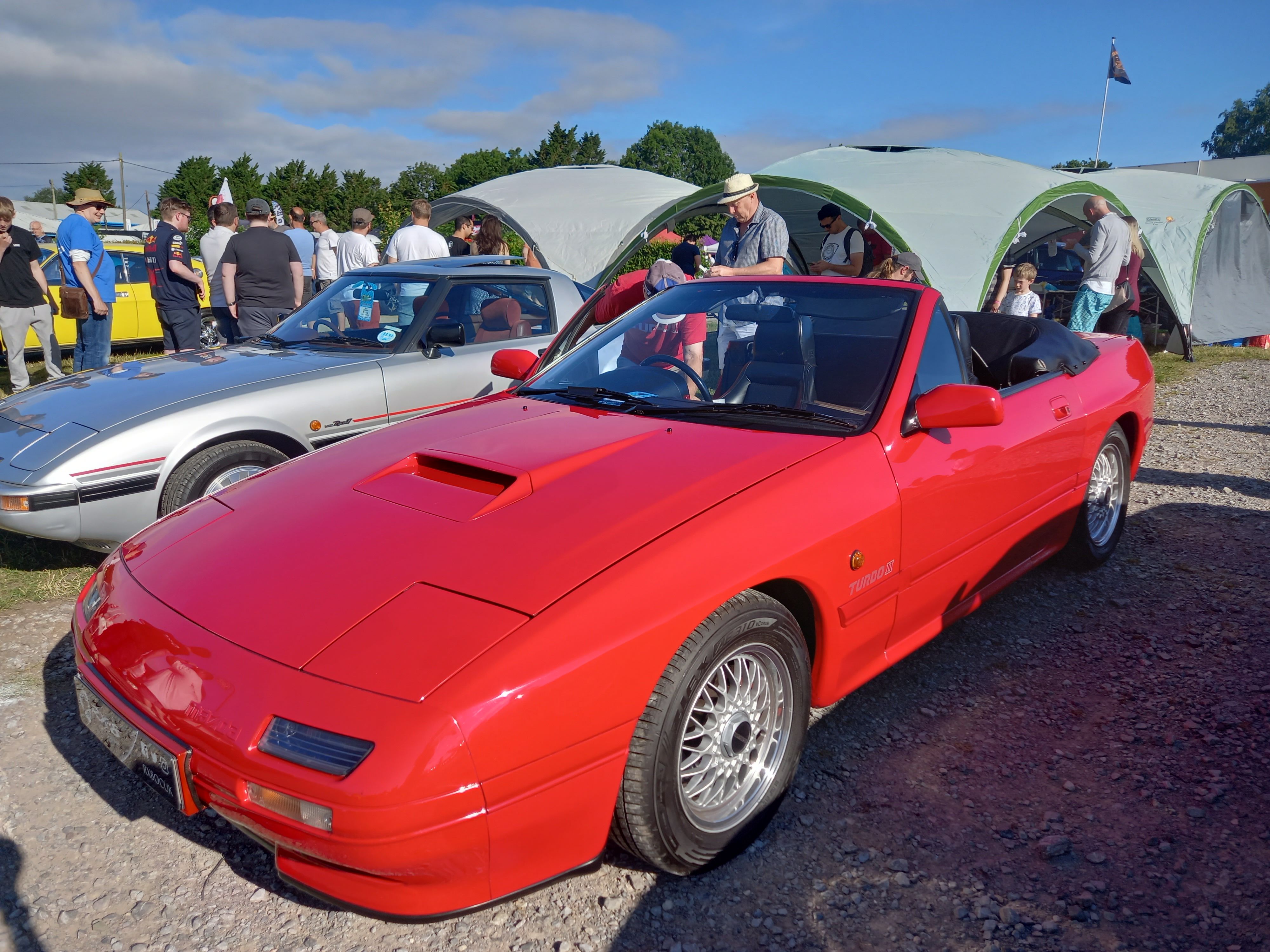 Front three-quarter view of a red second-generation Mazda RX-7 Turbo II convertible displayed outdoors on gravel at a classic car event.