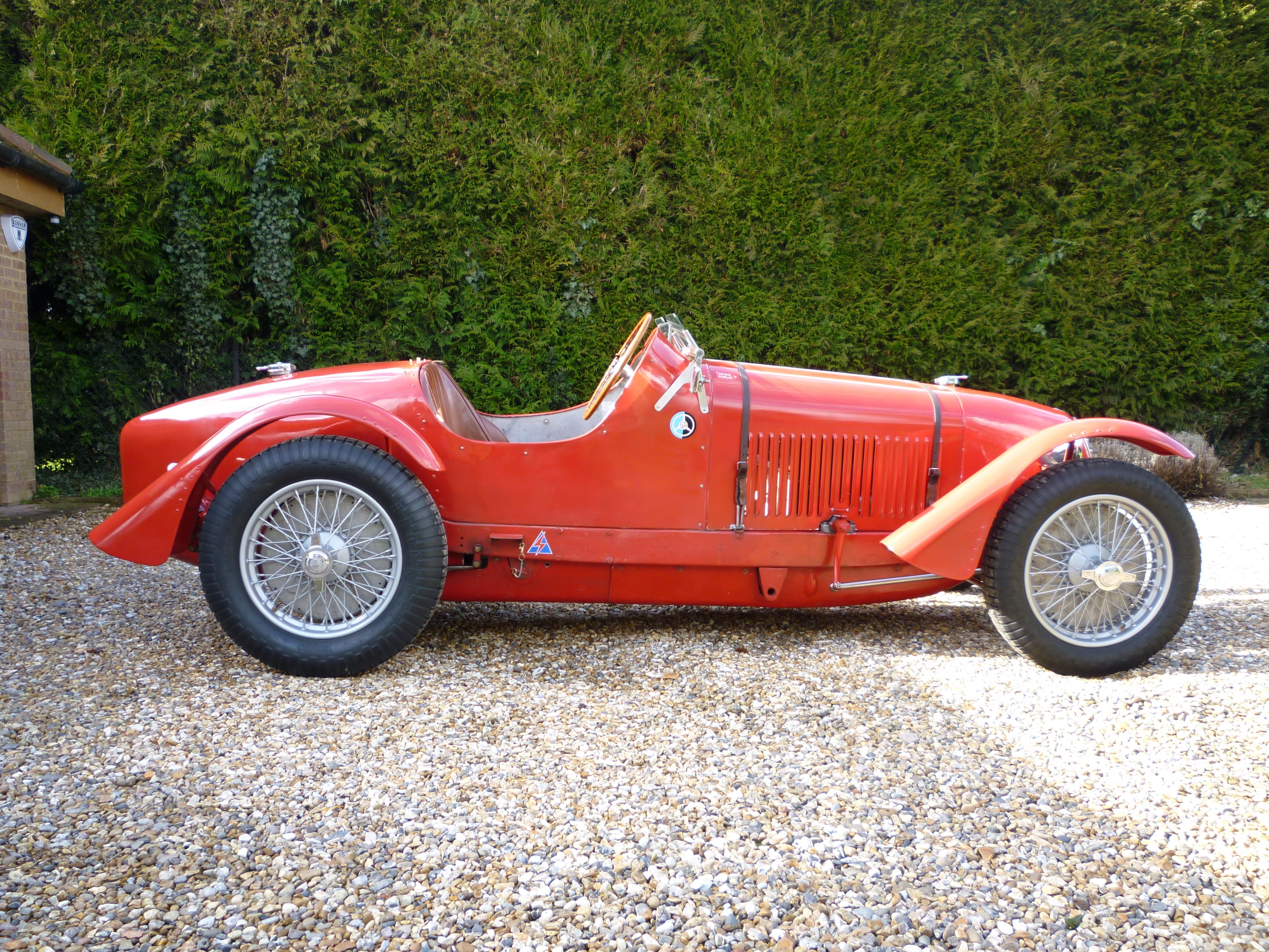 Side-profile view of a classic red open-top vintage sports car with wire-spoke wheels parked on a gravel driveway in front of a green hedge.