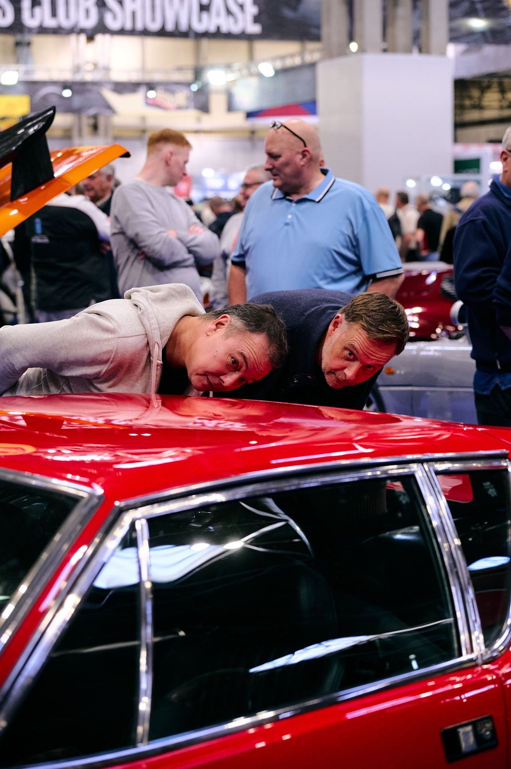 Two classic car enthusiasts leaning over to look inside a vintage red car at a crowded indoor automotive exhibition.