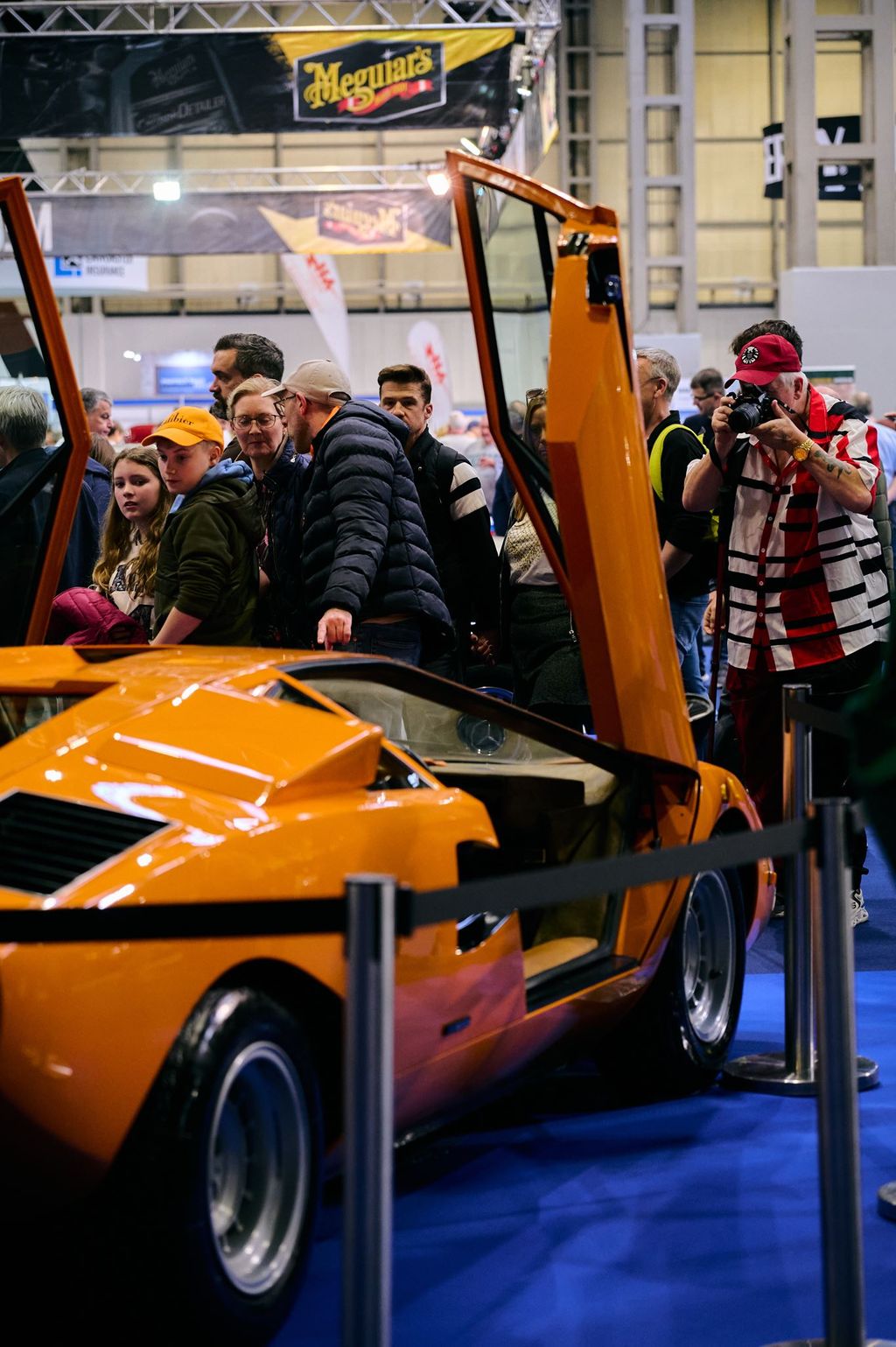 Orange Lamborghini Countach LP400 Periscopio with scissor doors open on display at a classic car show exhibition with crowds and Meguiar's banners.