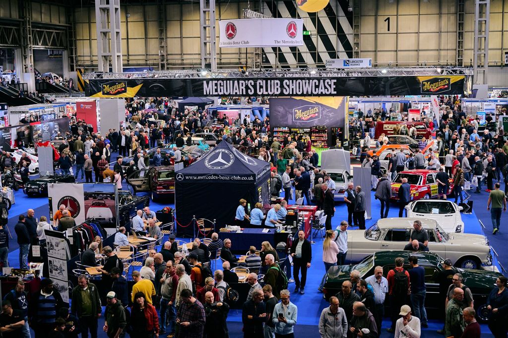 Wide shot of a busy classic car show and club exhibition hall packed with automotive enthusiasts, classic vehicles, and commercial display stands.