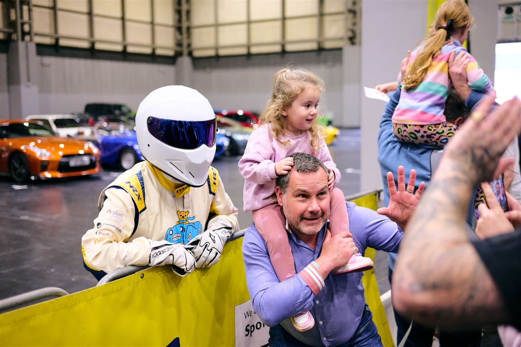 A man with a young child on his shoulders poses next to a person dressed as the Top Gear character The Stig, wearing a white racing helmet and suit, behind a yellow barrier at an indoor car show.