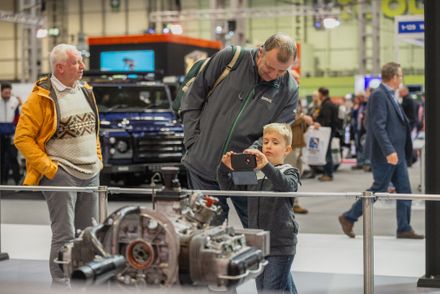A young boy takes a photo on a smartphone of an air-cooled classic car engine on display, while an adult stands beside him looking on, at a classic motor show exhibition hall.