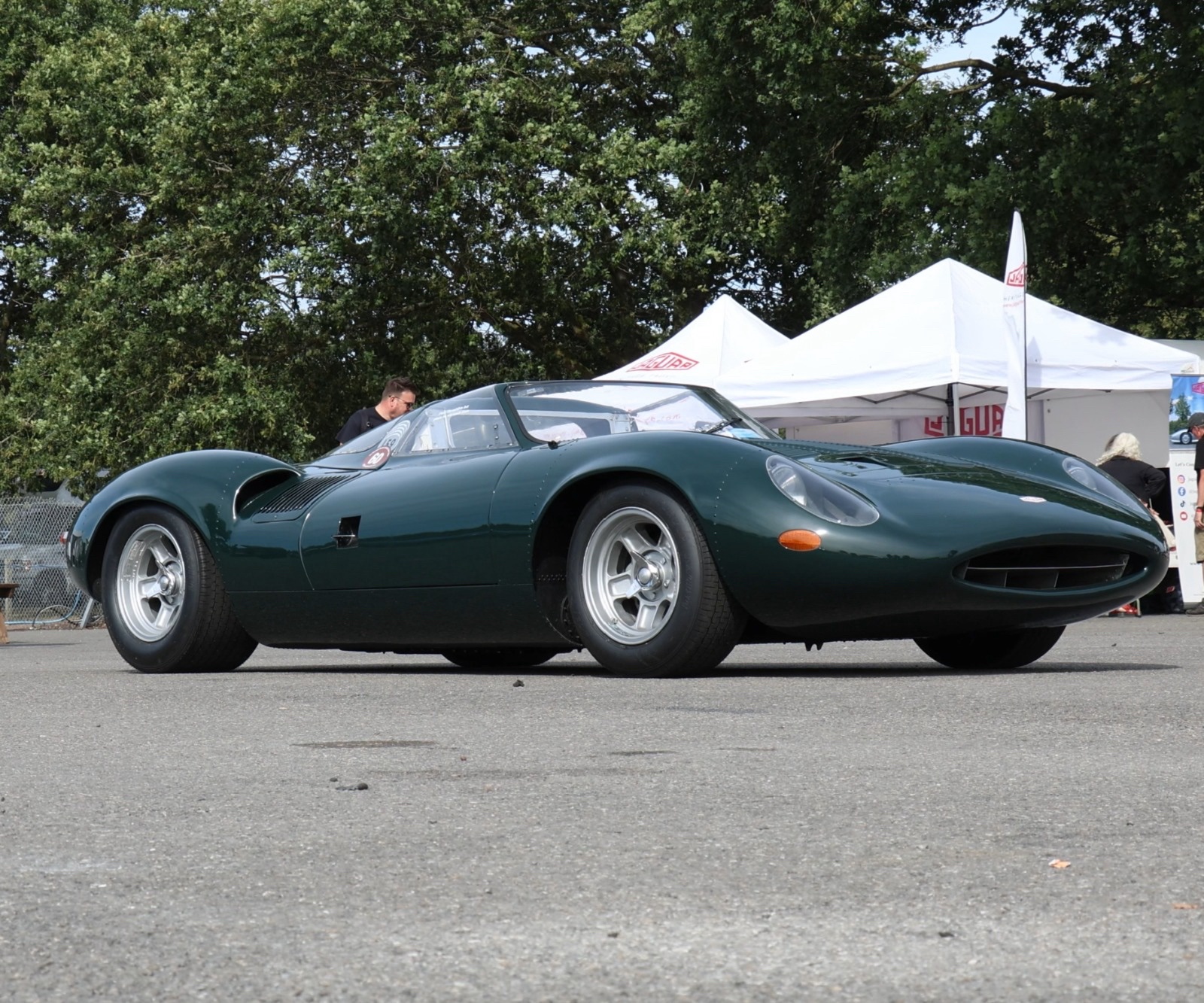 Low-angle view of a British racing green Jaguar XJ13 on tarmac, with white Jaguar gazebos and trees behind.