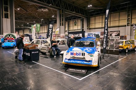 A wide shot of the 6R4.net stand at the Classic Motor Show, featuring a prominent blue and white MG Metro 6R4 rally car in the foreground. Enthusiasts explore the display, which includes car shells and memorabilia, within a large exhibition hall.