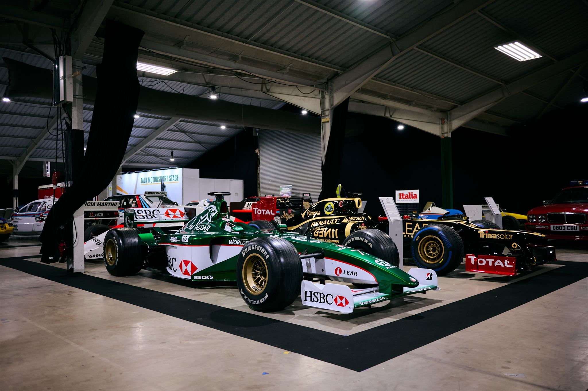 Classic Formula 1 cars including a green Jaguar Racing F1 car and a black Lotus Renault on display in a hangar.