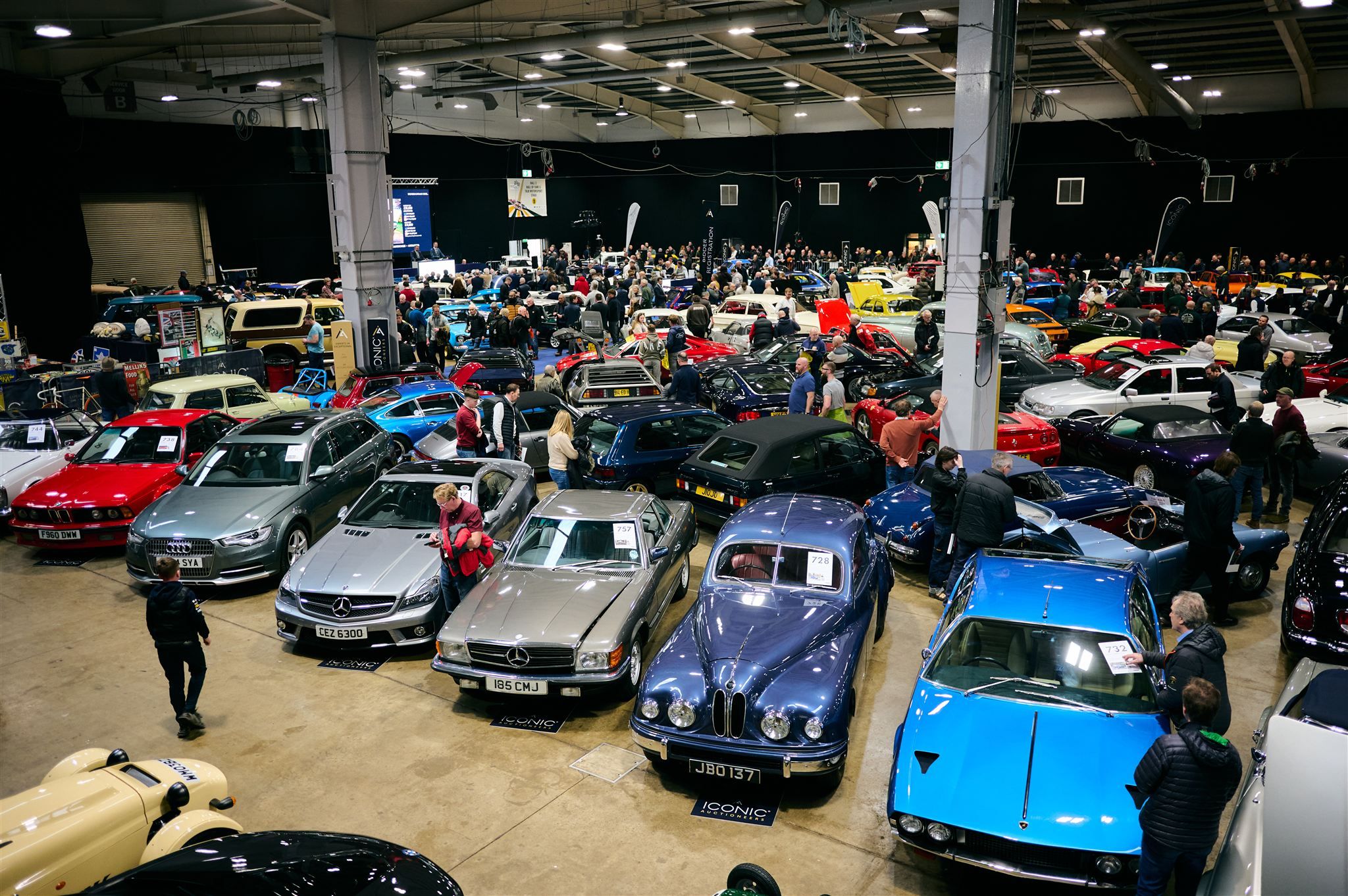 Large indoor classic car auction featuring a blue Bristol 401, a Lamborghini Jarama, and various Mercedes-Benz models by Iconic Auctioneers.