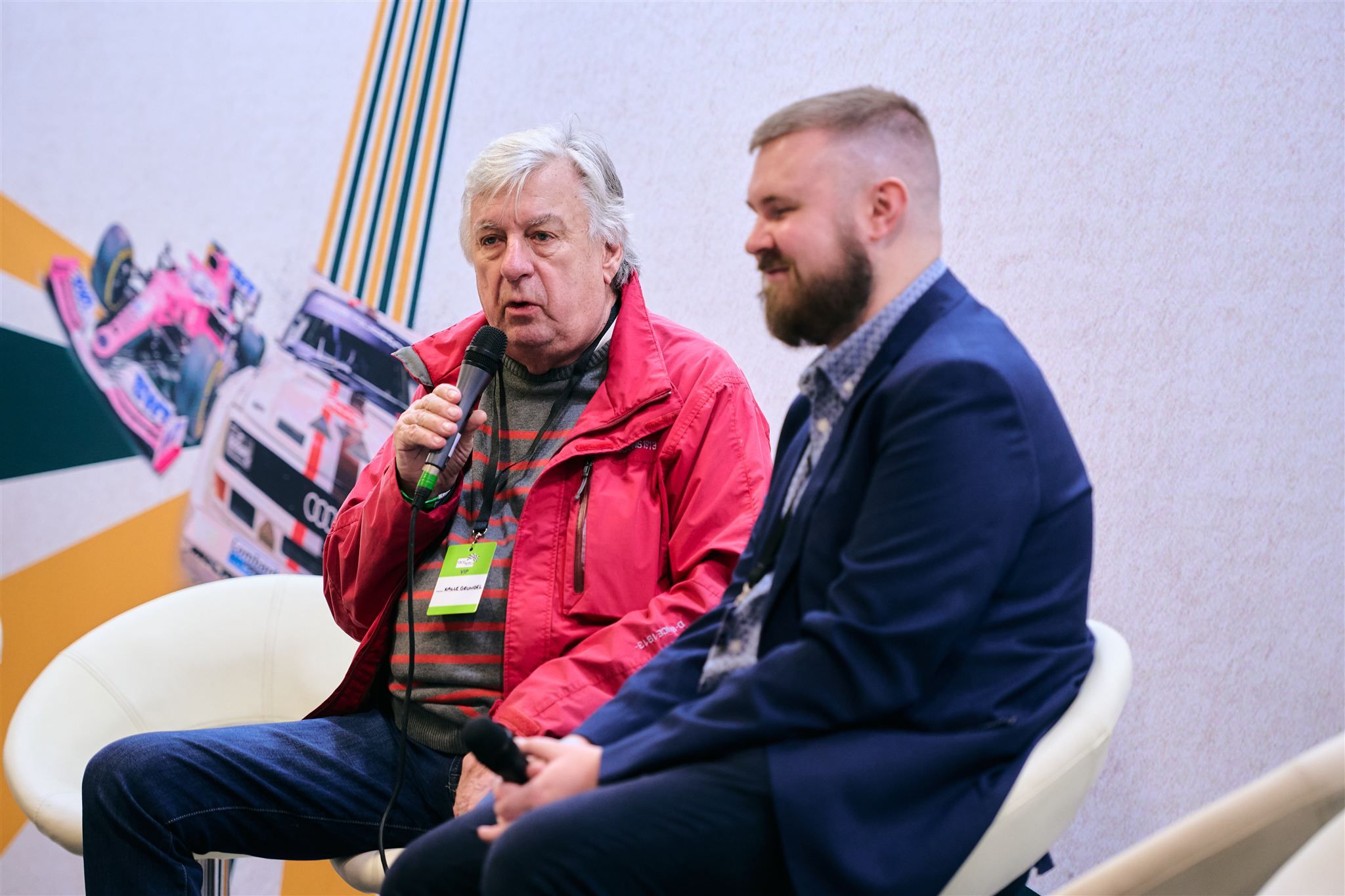 Rally legend Kalle Grundel in a red jacket sharing stories during a live Q&A at a motorsport event.