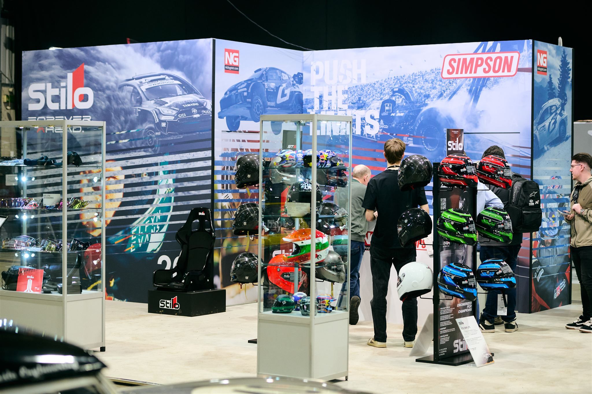 Stilo and Simpson motorsport safety equipment display featuring racing helmets, a racing seat, and gear at a professional trade show.