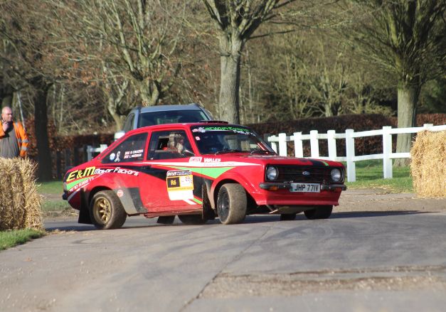 Red and black Ford Escort MK2 rally car driven by Gavin Crozier taking a sharp turn on a tarmac rally stage.