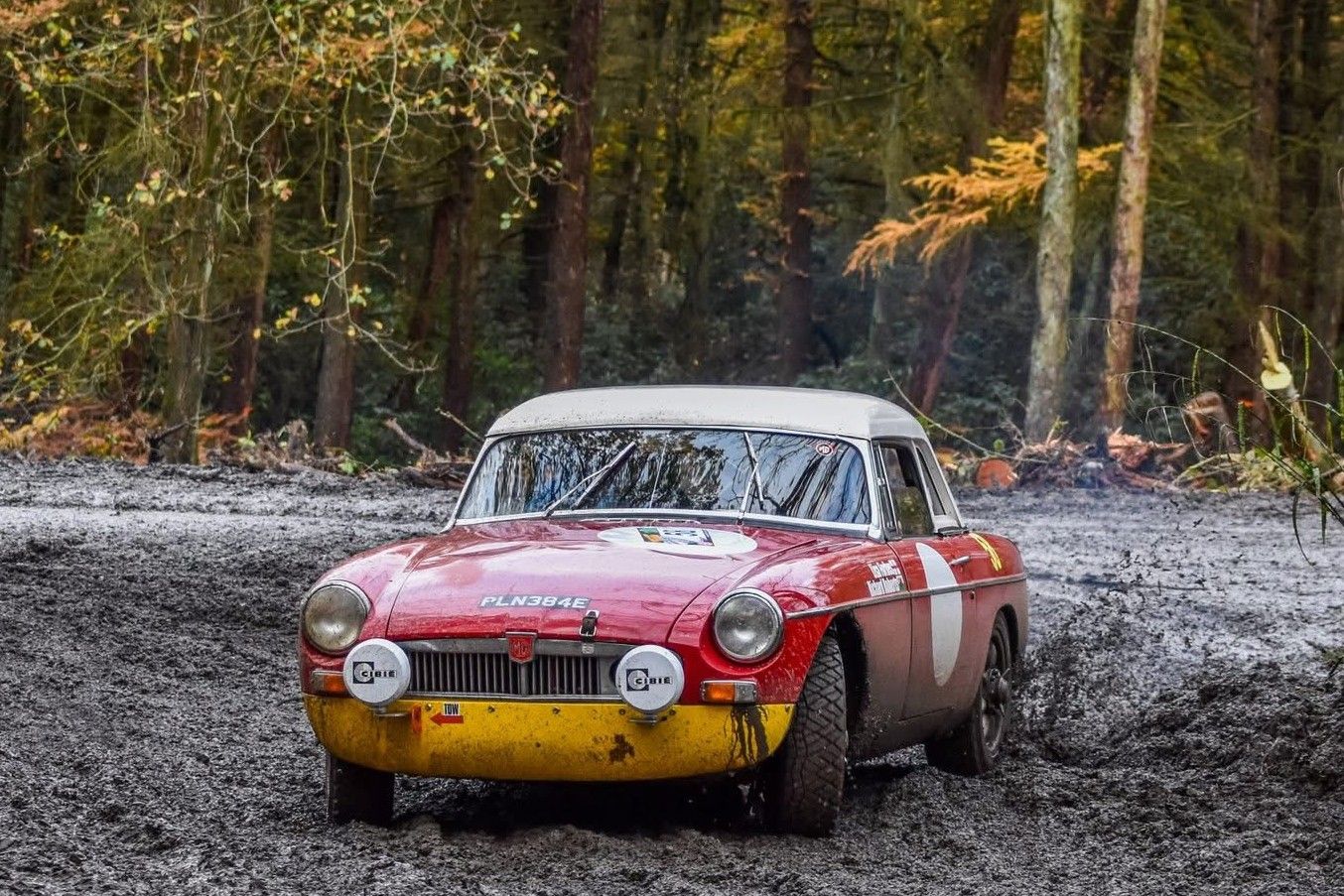 Classic red MGB Roadster rally car with a hardtop and yellow front valance driving through deep mud in a forest.