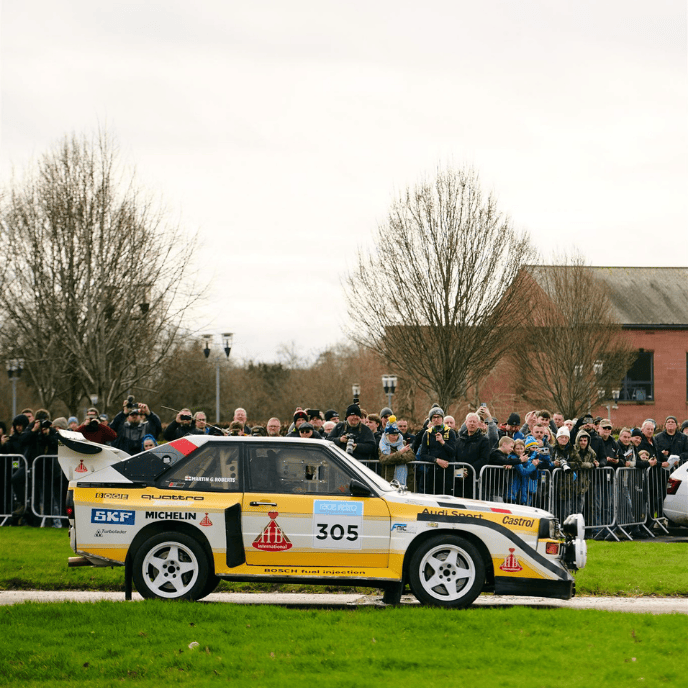Iconic yellow and white Audi Sport Quattro S1 rally car, number 305, driving past a crowd of spectators.