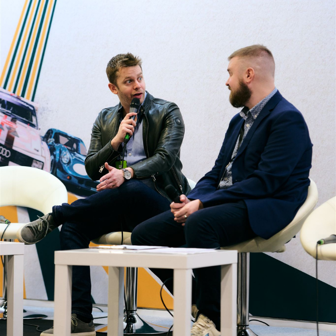 Alex Brundle and Chaz Draycott on stage at a motorsport event sitting in white chairs and speaking into microphones.