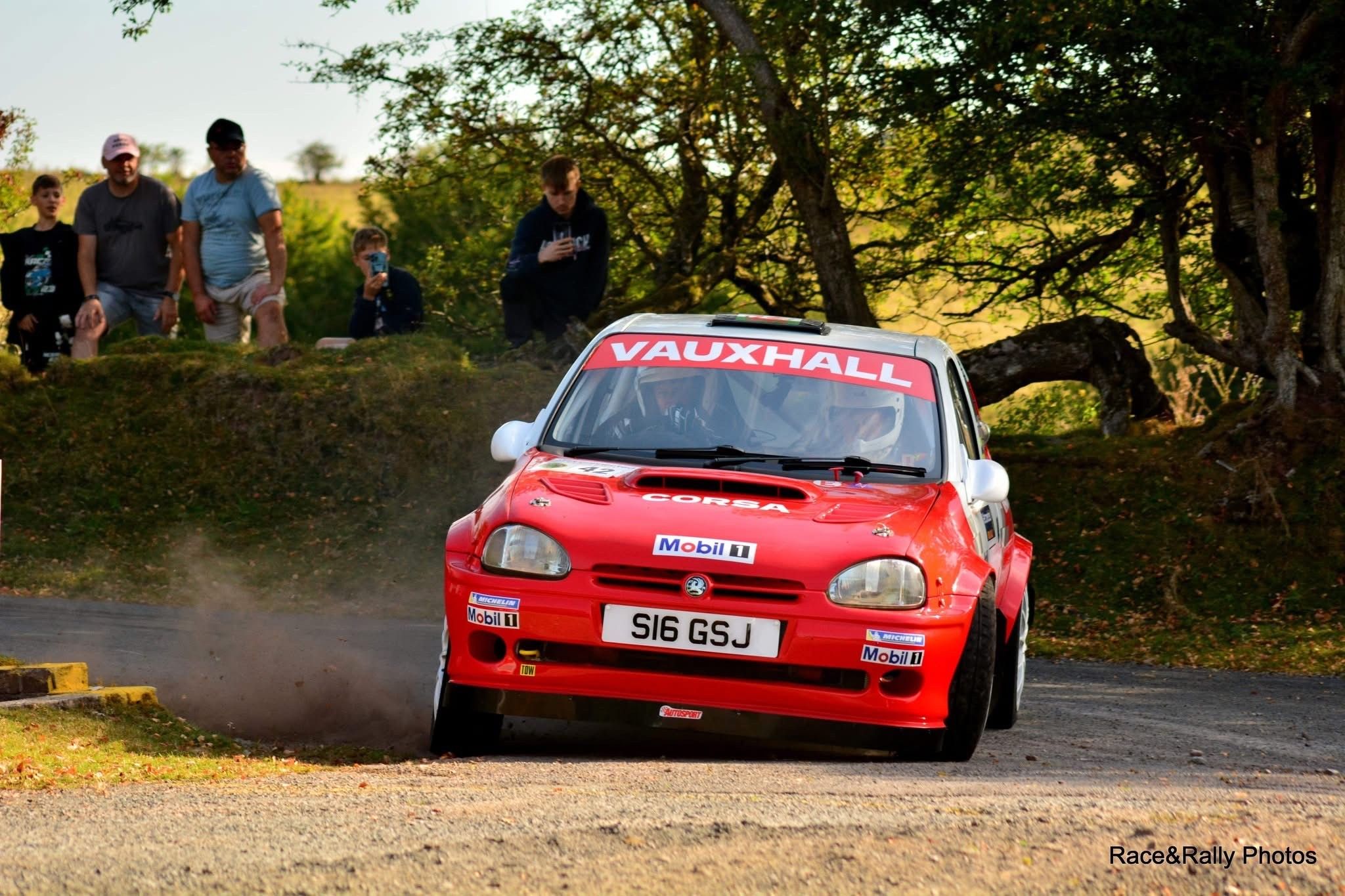 Red and white Vauxhall Corsa Kit Car driven by Aaron Jones navigating a dusty corner on an asphalt rally course.
