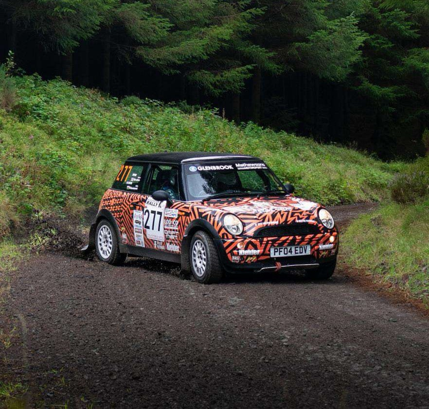Mini Cooper rally car with orange and black tiger stripe livery racing on a gravel forest track.