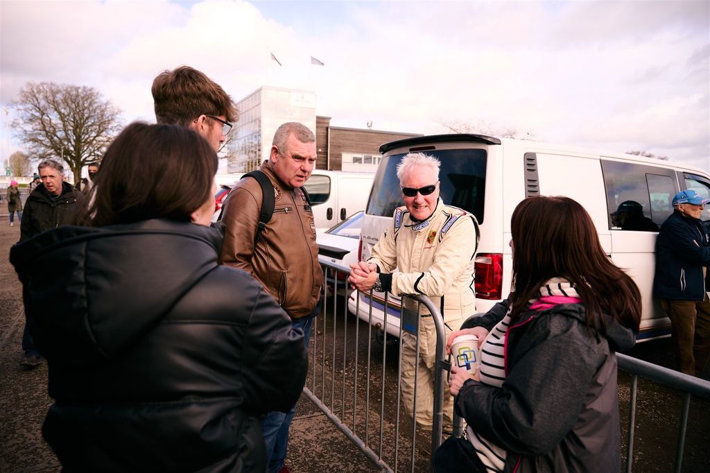  A man in a white racing jumpsuit and sunglasses talking to a group of people at a motorsport event.