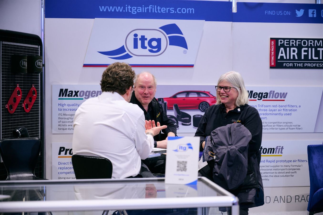 Three people talking at an ITG Air Filters trade show booth with a red Audi R8 in the background.
