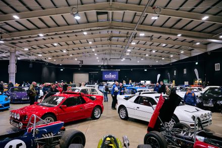 A wide variety of classic and modern performance cars, including a red Ford RS200 and a white Lotus Esprit, on display at an Iconic Auctioneers event in a large indoor showroom.