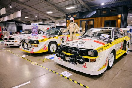 Three vintage Audi Quattro S1 rally cars with iconic yellow and white Audi Sport liveries on display at a motorsport exhibition featuring a racing driver mannequin.