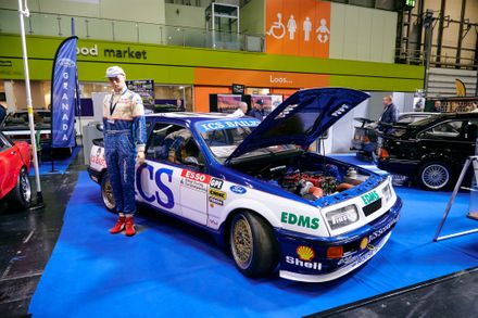Classic blue and white Ford Sierra RS500 Cosworth touring car with its hood open, displayed alongside a racing driver mannequin at an indoor motorsport exhibition.