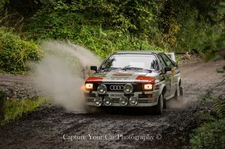 Audi Quattro Group 4 rally car splashing through a muddy puddle on a forest dirt track.