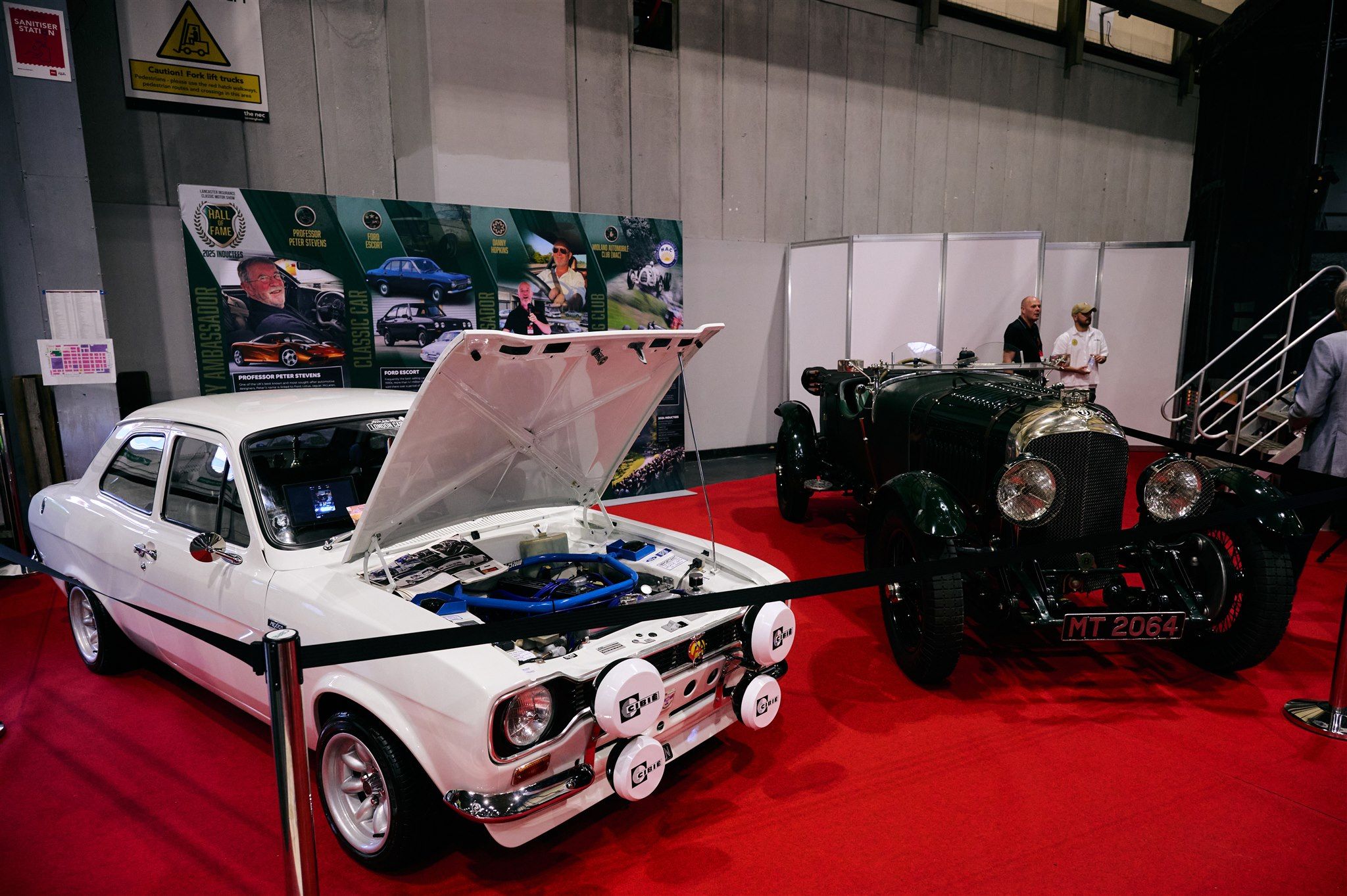 Modified white Ford Escort Mk1 with Cibie spotlights and open bonnet displayed next to a vintage Bentley on a red carpet at a car show.