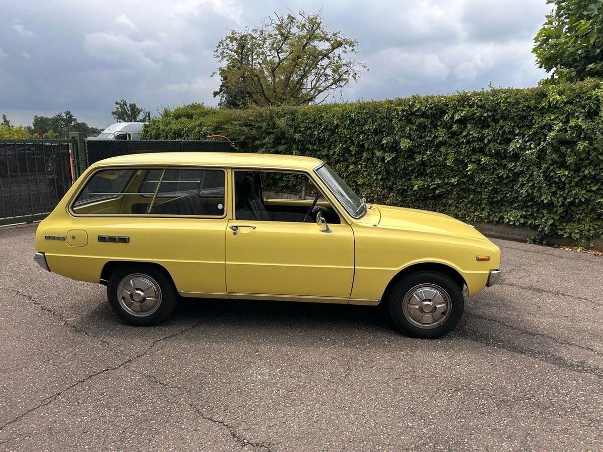 Vintage yellow Mazda 800 Estate parked on asphalt against a tall green hedge.