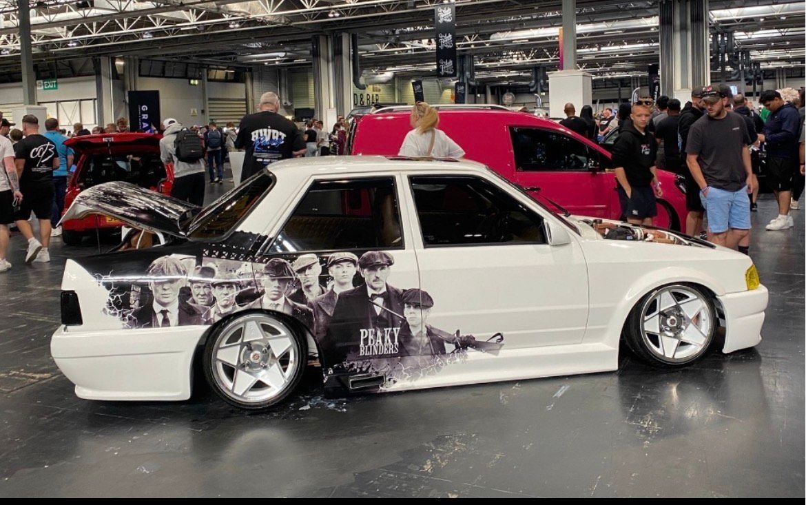 Custom white Ford Orion sedan with Peaky Blinders themed side-body wrap and deep-dish star wheels at an indoor car show.