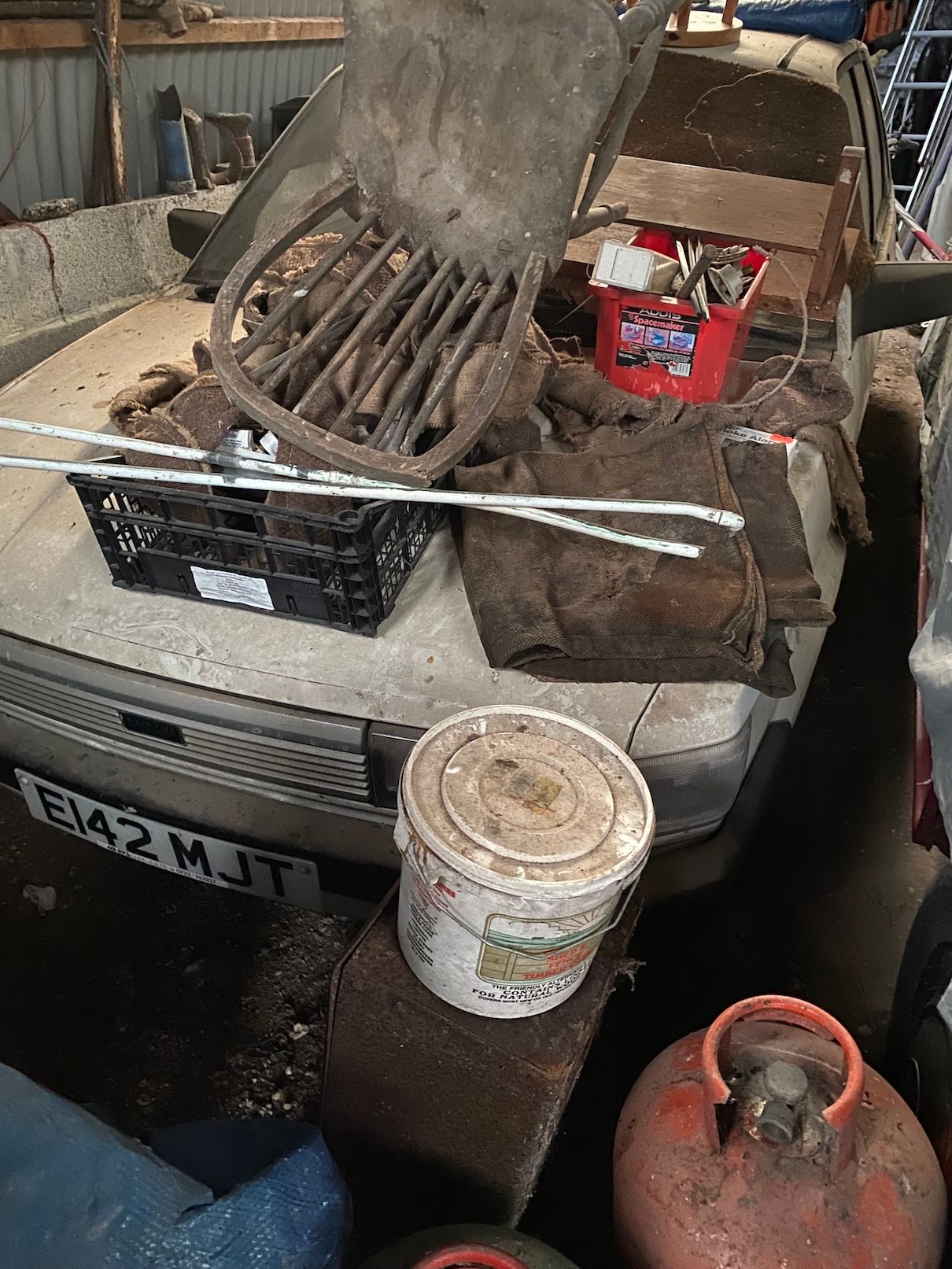 Dusty white Austin Maestro project car covered in storage items and burlap sacks in a garage.