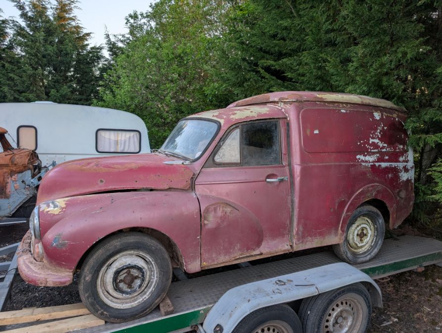 Rusty red Morris Minor panel van project being transported on a car trailer.