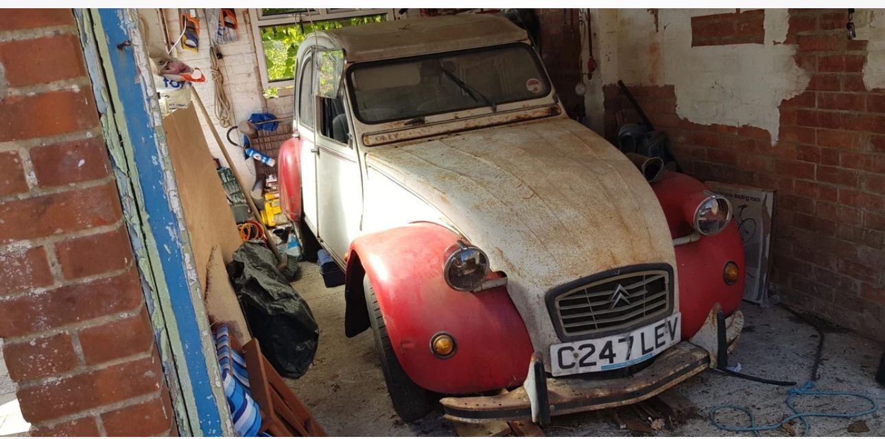 Red and white Citroën 2CV "Dolly" barn find with a rusted bonnet stored in a cluttered brick garage.
