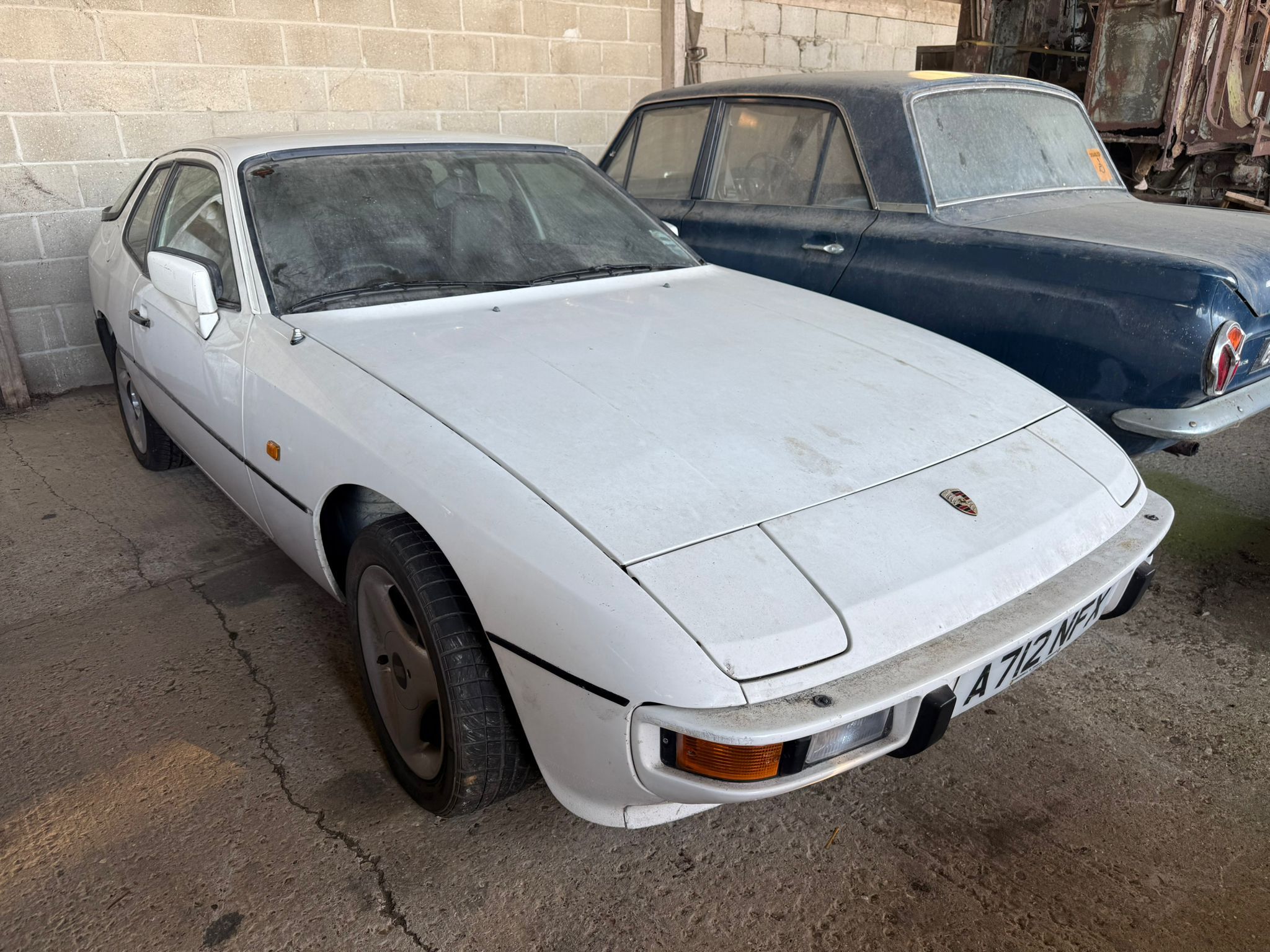 White Porsche 924 project car with popped-up headlights and moss growth in an outdoor barn find setting.