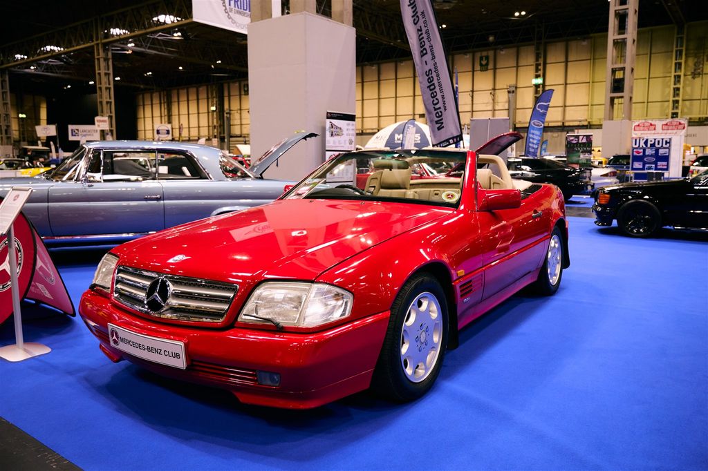Red Mercedes-Benz SL (R129) convertible classic car on display at the Mercedes-Benz Club stand during an indoor automotive show.