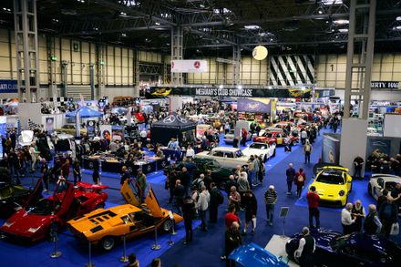 A high-angle view of a crowded indoor classic car show featuring a yellow Porsche, orange and red Lamborghini Countachs, and various vintage cars at the Meguiar's Club Showcase.