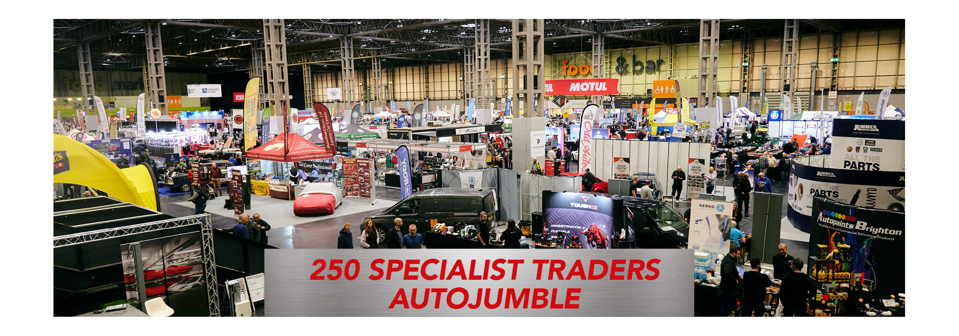 A busy indoor classic car autojumble at a large exhibition hall featuring over 250 specialist traders and automotive parts stalls.