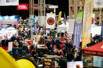 Large crowd of enthusiasts at the Practical Classics Restoration Show, featuring vintage MG cars, insurance vendor banners, and automotive club displays in a busy exhibition hall.