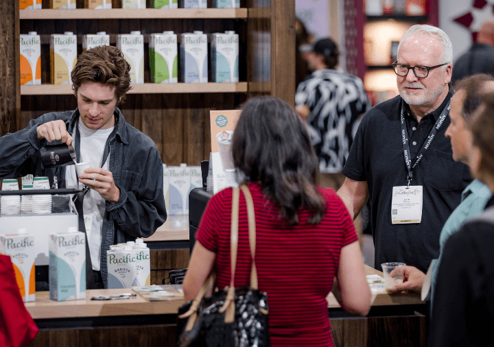 Pacific Barista Series exhibitors talking to attendee and preparing a sample on the show floor at Coffee Fest
